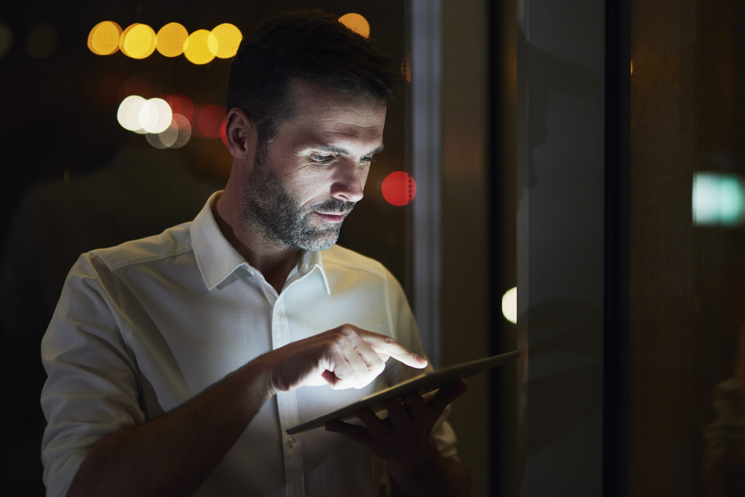 Profesional con camisa formal usando una tablet en un entorno corporativo nocturno, con luces desenfocadas al fondo, representando tecnología, activos digitales y seguimiento financiero.