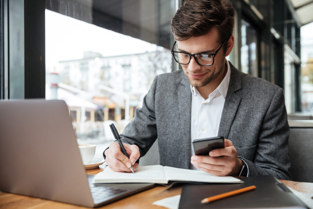 Homem profissional usando notebook e smartphone enquanto faz anotações, representando análise financeira, câmbio e estruturas internacionais.
