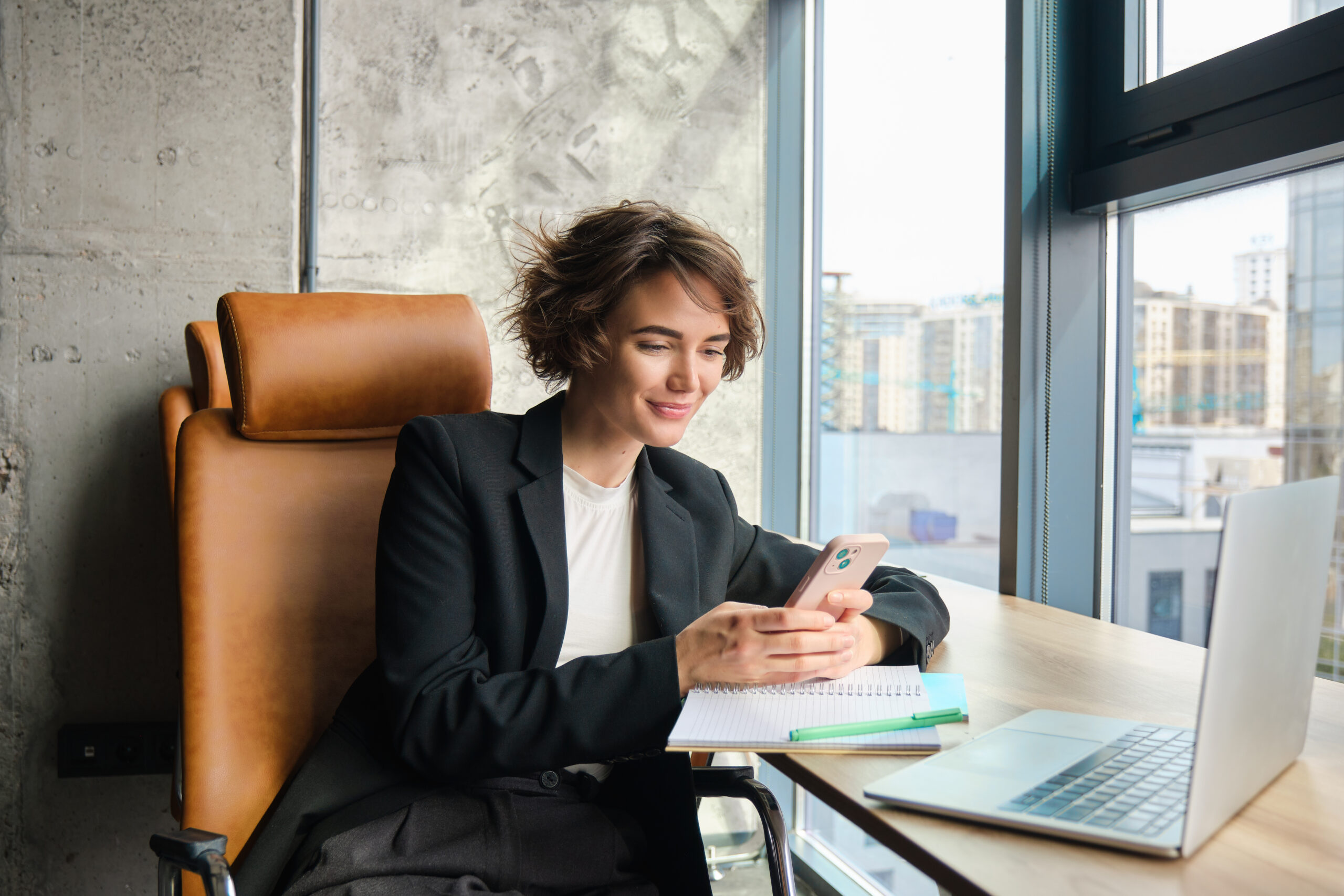 Mulher profissional usando smartphone e notebook em escritório, representando análise de investimentos digitais e acesso a informações financeiras.